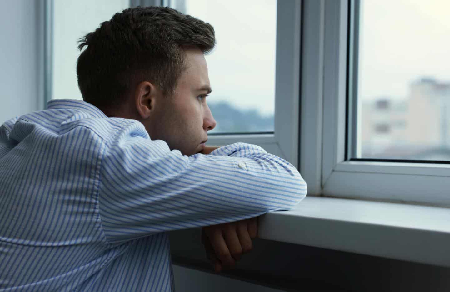 Man looking out a window with a depressed expression.